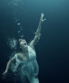 a woman in a blue dress under water with her arms raised up to the sky