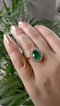 a woman's hand holding an emerald and diamond ring in front of a potted plant