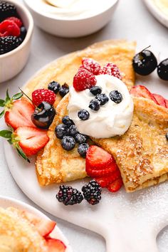 a white plate topped with french toast covered in berries and whipped cream next to bowls of fruit