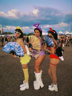 three women dressed in costumes at an outdoor music festival, one holding a fan and the other wearing knee high boots