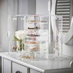 a clear jewelry organizer on top of a white dresser next to a flower and mirror