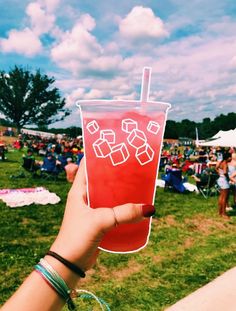 a person holding up a drink in front of an outdoor event with people sitting on the grass