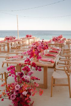 a table set up with pink flowers and place settings for an outdoor dinner on the beach