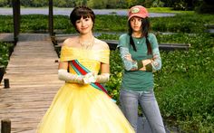 two young women dressed in costumes standing on a wooden bridge near water and grass, one wearing gloves