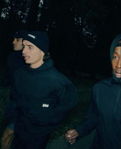 two men in black hoodies are walking through the woods at night with trees behind them