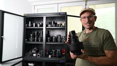 a man is holding a camera in front of a cabinet with many cameras on it