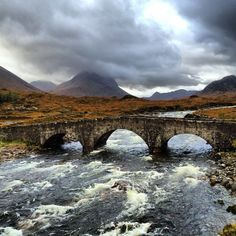 a stone bridge over a river with mountains in the background and cloudy skies above it