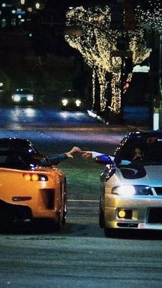 two cars parked next to each other on a street at night with lights in the background