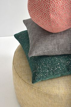 four pillows stacked on top of each other in different colors and patterns, all sitting on a round ottoman