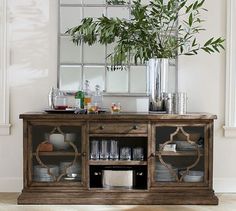 a wooden cabinet with plates and cups on it in front of a large mirror, filled with greenery