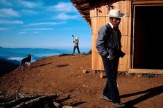 a man standing on top of a dirt hill next to a wooden building with a cow in the background