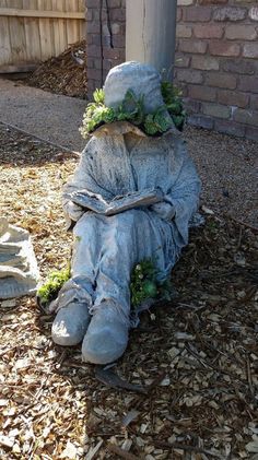 a statue sitting on the ground next to a brick wall and tree trunk with plants growing out of it