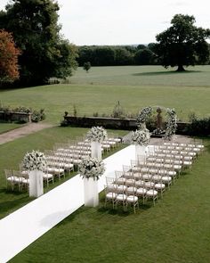 an outdoor ceremony setup with white chairs and floral arrangements on the aisle, in front of a field