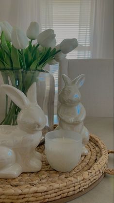 some white flowers are in a glass vase on a wicker tray with bunny figurines