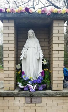 a statue of the virgin mary surrounded by flowers in a brick shrine with people sitting nearby