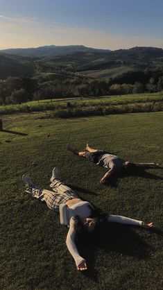 two people laying on the grass in front of a field with hills and rolling hills behind them