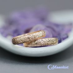 two gold wedding bands sitting on top of a white plate with purple flowers in the background