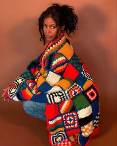 a woman wearing a multicolored crocheted sweater and jeans crouching on the ground