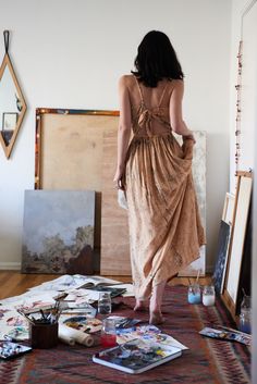 a woman in a brown dress is looking at pictures on the floor with easels and paintings behind her