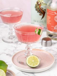 two glasses filled with pink lemonade and limes on a marble counter top next to bottles of gin