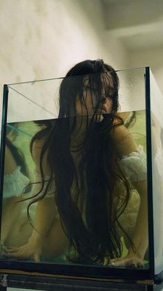 a woman with long hair sitting in front of a glass box on top of a shelf