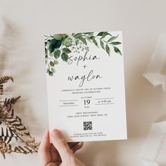 a person holding up a wedding card with greenery on it