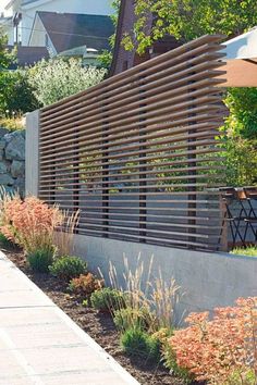 a wooden slatted fence next to a sidewalk with plants and flowers in the foreground