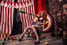 a woman with red hair sitting on the floor in front of a circus tent holding a green bat