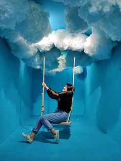 a man sitting on a swing in the middle of a room with clouds above him