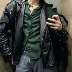 a man in a black leather jacket taking a selfie with his cell phone while standing next to a wall