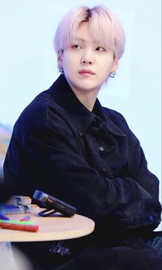 a young man with pink hair sitting at a table and looking off to the side