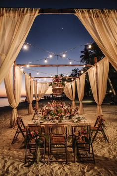 an outdoor dining set up on the beach at night with string lights and draping