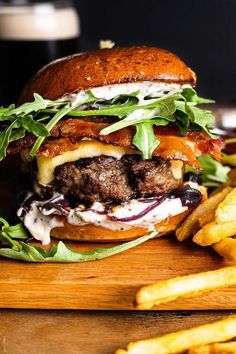 a burger with lettuce, bacon and cheese on a cutting board next to french fries