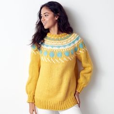 a woman standing in front of a white wall wearing a yellow sweater and white pants