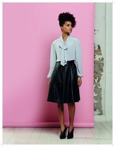 a woman standing in front of a pink wall wearing a white shirt and black leather skirt