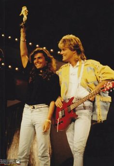 two people standing on stage with guitars and one person holding an object in the air