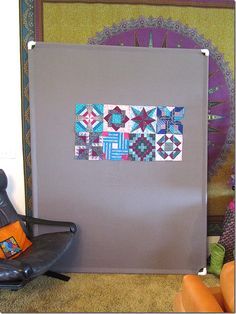 a chair sitting in front of a gray wall with patchwork on it and an orange pillow next to it