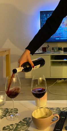 a person pouring red wine into two glasses on a table with a television in the background