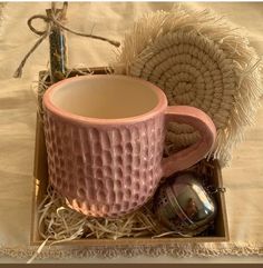 a pink coffee cup sitting in a box on top of a bed next to a pillow