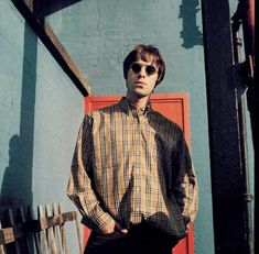 a man standing in front of a blue wall wearing sunglasses and a plaid shirt with his hands in his pockets