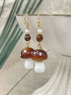 Handmade, adorable lampwork glass mushroom earrings. These earrings feature a charming brown glass mushroom bead with delicate white spots. The brown glass mushroom bead is skillfully paired with gold-plated wires and complemented by matching brown Czech glass beads. These trendy mushroom earrings are perfect for adding a touch of whimsy to your everyday outfits.  They would make the perfect gift for nature enthusiasts or anyone who loves unique accessories. For your convenience and peace of min Mushroom Bead Earrings, White Mushroom Design Earrings For Gift, White Earrings With Mushroom Design As Gift, White Mushroom Design Earrings As Gift, Whimsical White Mushroom Design Earrings, Unique White Jewelry With Mushroom Design, Unique White Mushroom Design Jewelry, White Dangle Earrings With Mushroom Design, White Mushroom-shaped Jewelry With Mushroom Design