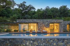 an outdoor swimming pool surrounded by stone walls and trees at dusk with the lights on