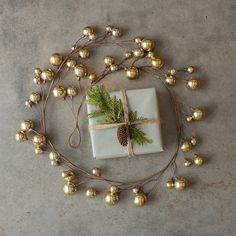 a present wrapped in white paper surrounded by gold balls and greenery with a pine cone on top