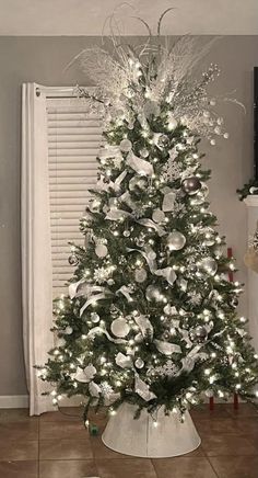 a white christmas tree with silver ornaments and lights in a living room next to a window