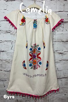 Precious Mexican dress for girls with fun colorful poms around dropped shoulder sleeve and hem line. Dress has fine thread frontal embroidery. Color selection is based on pom-pom color. Please note that embroidery pattern and thread colors may not be identical as pictured. Made with 100% raw cotton (manta). COLOR AND SIZE AVAILABILITY VARY. BE SURE TO SELECT COLOR OPTIONS FOR THE SIZE NEEDED. SIZE WIDTH LENGTH 6MTH 13 13 1 YR 14 15 2 T 18 19 4 T 18 22 6 YR 20 25 8 YR 20 26 10 YR 21 28 Measuremen Mexican Dresses, Shoulder Sleeve, Floral Embroidery, Head Wraps, Color Options, Embroidery Patterns, Pom Pom, Cover Up, Girls Dresses