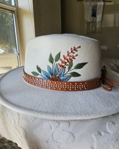 a white hat with flowers on it sitting on top of a table next to a window