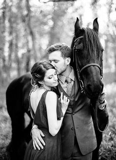 a man and woman standing next to a horse in the woods with their head close to each other