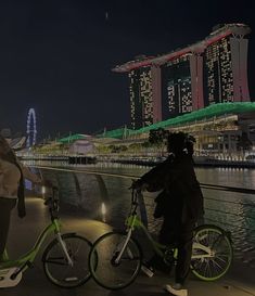 two people standing next to each other on bikes near the water and buildings at night