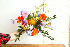 a woman holding a bouquet of colorful flowers on top of a wooden table in front of a white brick wall