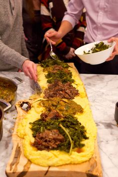 two people are serving themselves food on a long wooden platter with greens and meat